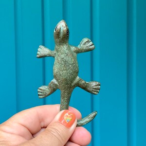 May include: A green ceramic lizard figurine with a textured surface. The lizard is standing on its hind legs with its front legs outstretched. The lizard has a long tail that curls up behind it.