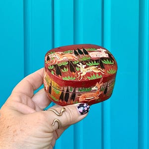 May include: A small, red, hand-painted box with a lid. The box features a detailed illustration of deer and foliage. The box is held in a hand against a bright blue background. The box is likely used for storing small items.