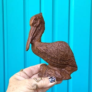 May include: A brown, carved wooden pelican sculpture. The bird is depicted in a resting pose, with detailed feather and beak carvings. The sculpture is held against a turquoise background.