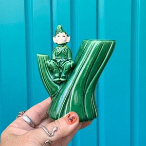 May include: A green ceramic planter shaped like a tree trunk with a small green and white ceramic gnome sitting on top.