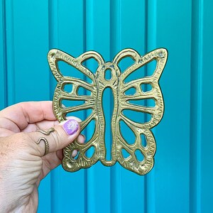 May include: A brass butterfly-shaped decorative object with intricate cut-out details. The butterfly is held against a bright turquoise background. The object has a golden hue and is likely made of metal.