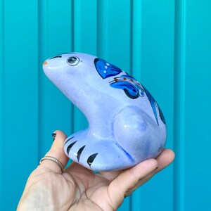 May include: A hand holds a blue and white ceramic frog figurine with painted floral details. The frog is sitting on its hind legs with its front legs outstretched.
