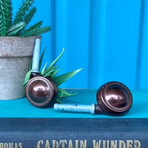 May include: Two brown metal furniture casters with silver metal stems. The casters are sitting on top of a black book with the title "Captain Wunder" and the author "Ald Thomas".