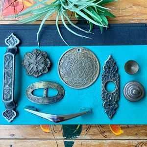May include: An assortment of antique metal hardware pieces, including handles, knobs, and decorative plates, in various shapes and finishes. The hardware is arranged on a teal surface, with a sprig of greenery at the top. The pieces are in shades of silver, bronze, and brass.