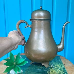 May include: A hammered brass coffee pot with a rounded lid, a curved spout, and a textured, woven-style handle. The pot features three small, decorative feet at the base and a warm, metallic finish throughout the body of the vessel.