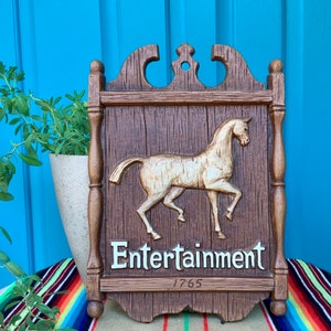 May include: A wooden sign with a carved horse and the words "Entertainment 1765". The sign is painted brown and has a white background for the text.
