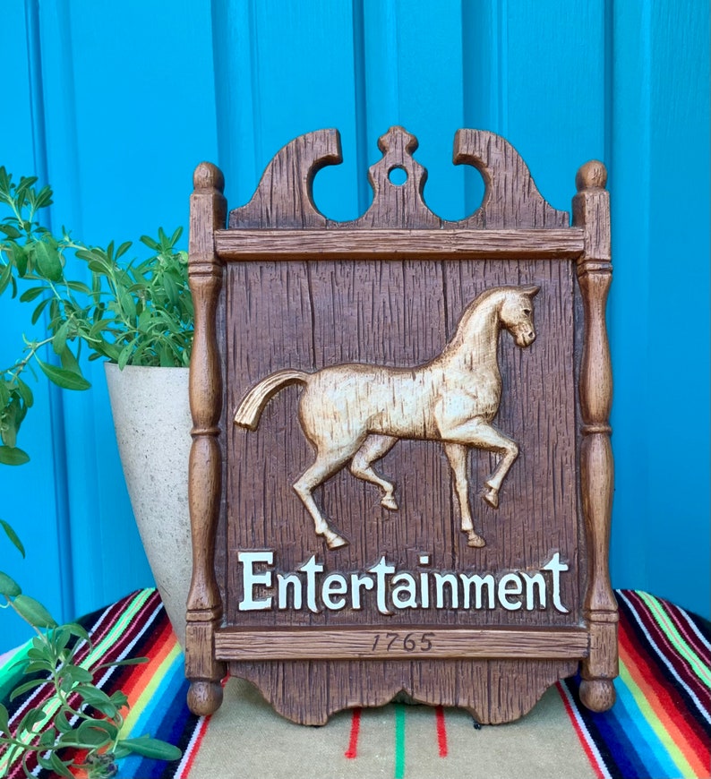 May include: A wooden sign with a carved horse and the words "Entertainment 1765". The sign is painted brown and has a white background for the text.