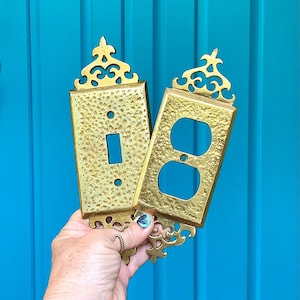 May include: Two ornate, gold-colored light switch and outlet covers. The covers have a textured surface and decorative filigree at the top and bottom. The background is a solid, bright blue.