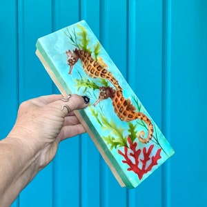 May include: A rectangular wooden box with a painted design of two seahorses swimming among coral and seaweed. The box has a light blue background with a white iridescent finish.
