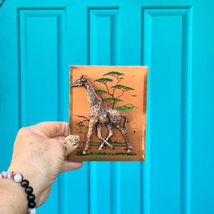 May include: A wooden plaque with a 3D sculpted giraffe in a brown and gold color scheme. The giraffe is walking through a green and brown landscape with trees. The plaque has a brown wood frame.
