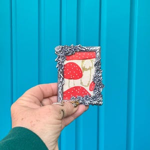 May include: A silver-toned picture frame with a floral design. The frame is holding a red and white patterned paper with a mushroom design.