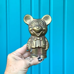 Op de afbeelding: Een gouden metalen beeldje van Mickey Mouse, gekleed in een pak en met een strikje. Het beeldje staat op een blauwe ondergrond.