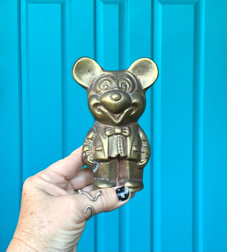 Op de afbeelding: Een gouden metalen beeldje van Mickey Mouse, gekleed in een pak en met een strikje. Het beeldje staat op een blauwe ondergrond.
