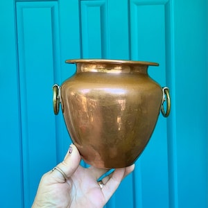 May include: A polished copper pot with a wide, rounded body and a flared rim. The vessel features two circular brass ring handles attached to the sides, providing a functional grip. The surface has a warm, metallic sheen reflecting light.