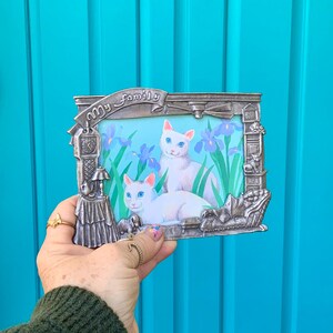 May include: A silver-colored picture frame with the words "My Family" on a banner. The frame holds a picture of two white cats with blue eyes and purple flowers. The frame has decorative elements including a clock, a lamp, and a small dog.
