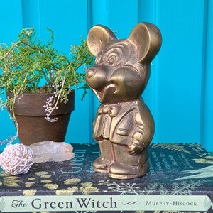 Op de afbeelding: Een messing beeldje van Mickey Mouse in smoking. Het beeldje staat op een stapel boeken met de titel "The Green Witch" zichtbaar.