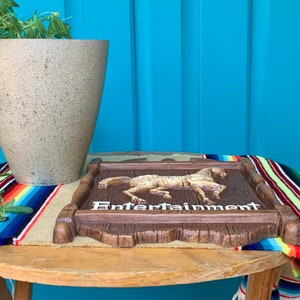 May include: A wooden sign with a carved horse and the word "Entertainment" in white letters. The sign is sitting on a table with a colorful blanket underneath.