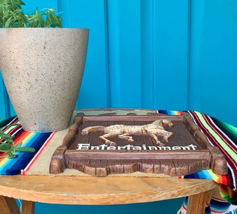 May include: A wooden sign with a carved horse and the word "Entertainment" in white letters. The sign is sitting on a table with a colorful blanket underneath.