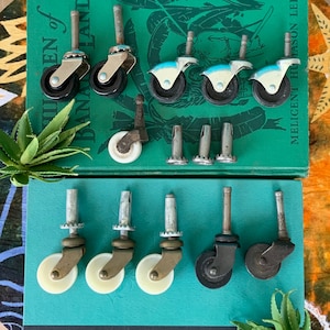 May include: Assortment of 11 furniture casters, including 4 black casters with white wheels, 3 white casters with brass accents, 3 white casters with silver accents, and 1 white caster with a silver accent. The casters are arranged on two books with the text "Children of Banana Land" and "Mellicent Hudson Lee".
