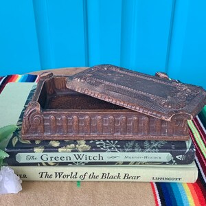 May include: A vintage wooden box with an ornate design and a hinged lid. The box is sitting on top of two books, one titled "The Green Witch" and the other titled "The World of the Black Bear".