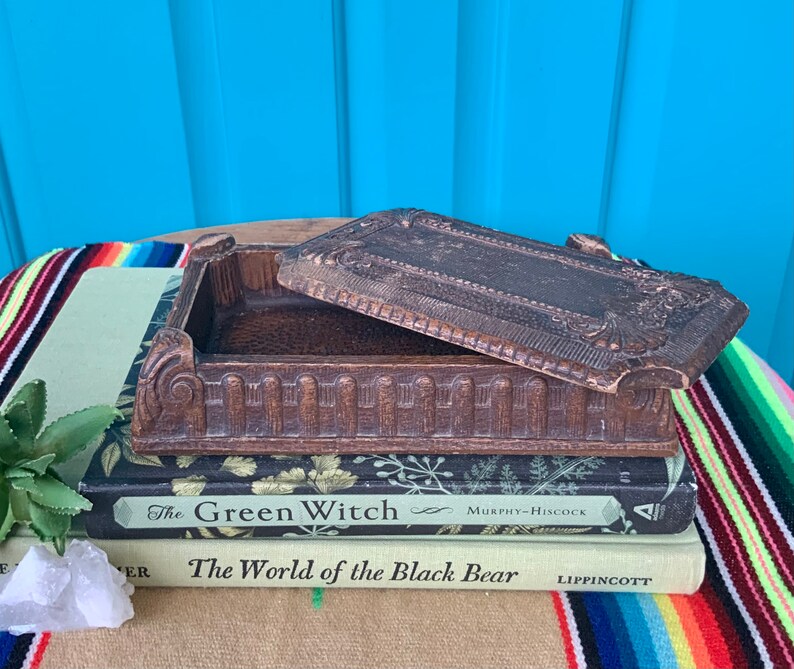 May include: A vintage wooden box with an ornate design and a hinged lid. The box is sitting on top of two books, one titled "The Green Witch" and the other titled "The World of the Black Bear".