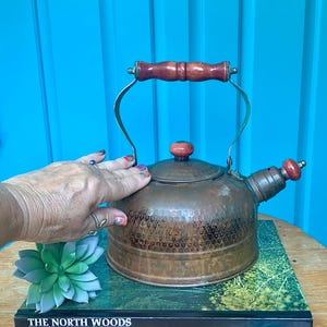 May include: A hammered copper tea kettle with a rounded body, a brass-toned metal handle, and a spout. The kettle features a wooden handle grip and a matching wooden knob on the lid. The surface shows a textured, dimpled pattern on the lower half.