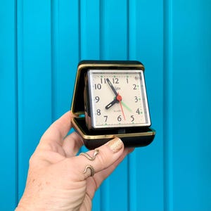 May include: A black and gold square-shaped alarm clock with a white face and black hands. The clock face has the word "Algin" and "QUART" printed on it. The clock is set to 10:10.