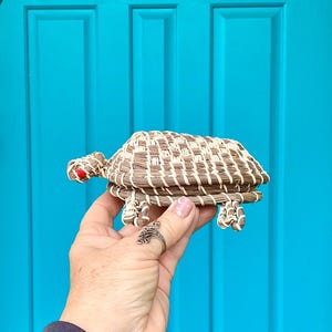 May include: A woven turtle figurine with a brown and cream-colored shell, red eyes, and four legs. The turtle is made of natural fibers and is held against a bright blue background.