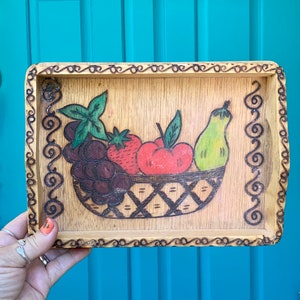 May include: A wooden tray with a hand-painted design of fruit in a basket. The fruit includes grapes, a pear, a strawberry, and an apple. The tray has a decorative border.