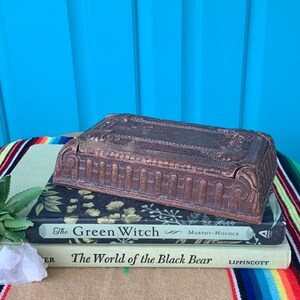 May include: A vintage wooden box with intricate carvings and a hinged lid. The box is sitting on top of two books, one titled "The Green Witch" and the other titled "The World of the Black Bear".