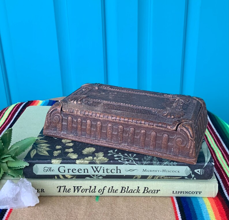 May include: A vintage wooden box with intricate carvings and a hinged lid. The box is sitting on top of two books, one titled "The Green Witch" and the other titled "The World of the Black Bear".