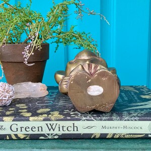 Op de afbeelding: Een messing beeldje van een aap die zit met zijn handen voor zich gevouwen. De aap ligt op zijn zij en heeft een gedetailleerd gezicht. Het beeldje staat op een boek met de titel "The Green Witch".