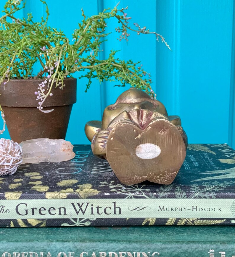 Op de afbeelding: Een messing beeldje van een aap die zit met zijn handen voor zich gevouwen. De aap ligt op zijn zij en heeft een gedetailleerd gezicht. Het beeldje staat op een boek met de titel "The Green Witch".