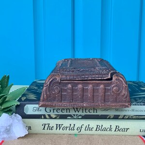 May include: A vintage wooden box with intricate carvings and a hinged lid. The box is sitting on top of two books, one titled "The Green Witch" and the other titled "The World of the Black Bear".