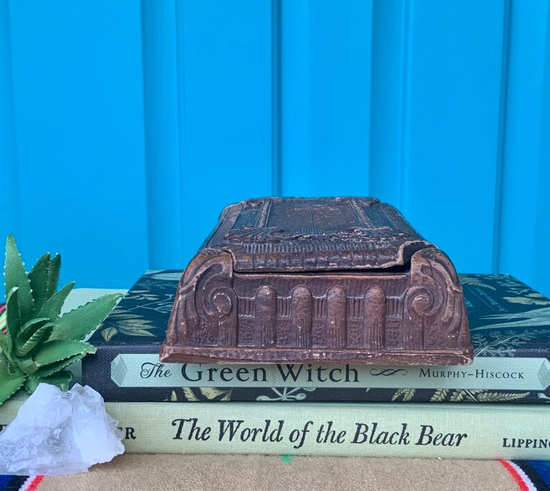 May include: A vintage wooden box with intricate carvings and a hinged lid. The box is sitting on top of two books, one titled "The Green Witch" and the other titled "The World of the Black Bear".