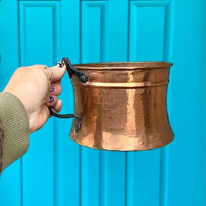 May include: A copper-colored metal pot with a curved body and a black handle. The pot has a wide opening and a slightly flared rim. The handle is attached to the side of the pot with rivets. The pot is held against a bright blue background.