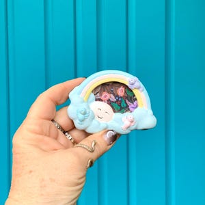 May include: A small, blue and pink ceramic picture frame shaped like a cloud with a rainbow and birds. The frame has a floral pattern inside.