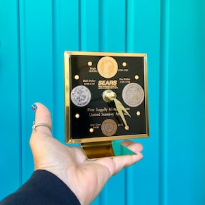May include: A black and gold clock featuring vintage coins, including an Eagle, Half-Dollar, and One Dollar coin. The clock face includes the text "SEARS" and "First Legally Minted in the United States of America."