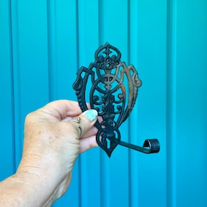 May include: Ornate black metal wall hook with a decorative filigree design. The hook has a curved arm and is held against a turquoise blue door. The metal shows some signs of rust.