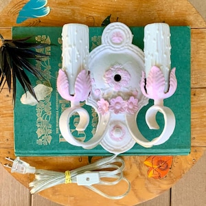 May include: A vintage white wall sconce with two candle holders, accented with pink floral and leaf details. The sconce is mounted on a green book. A power cord with a plug is visible.