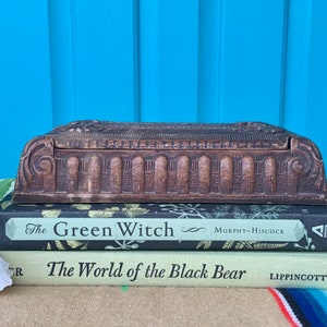 May include: A vintage wooden box with intricate carvings and a hinged lid. The box is sitting on top of two books, "The Green Witch" and "The World of the Black Bear".