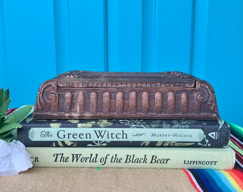 May include: A vintage wooden box with intricate carvings and a hinged lid. The box is sitting on top of two books, "The Green Witch" and "The World of the Black Bear".