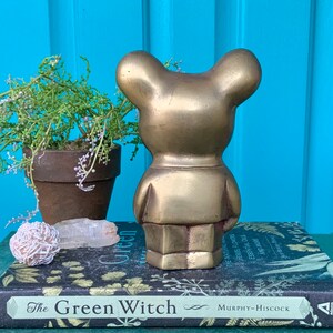 Op de afbeelding: Een gouden metalen berenbeeldje met een eenvoudig ontwerp. De beer staat op een stapel boeken met de titel "The Green Witch" zichtbaar.