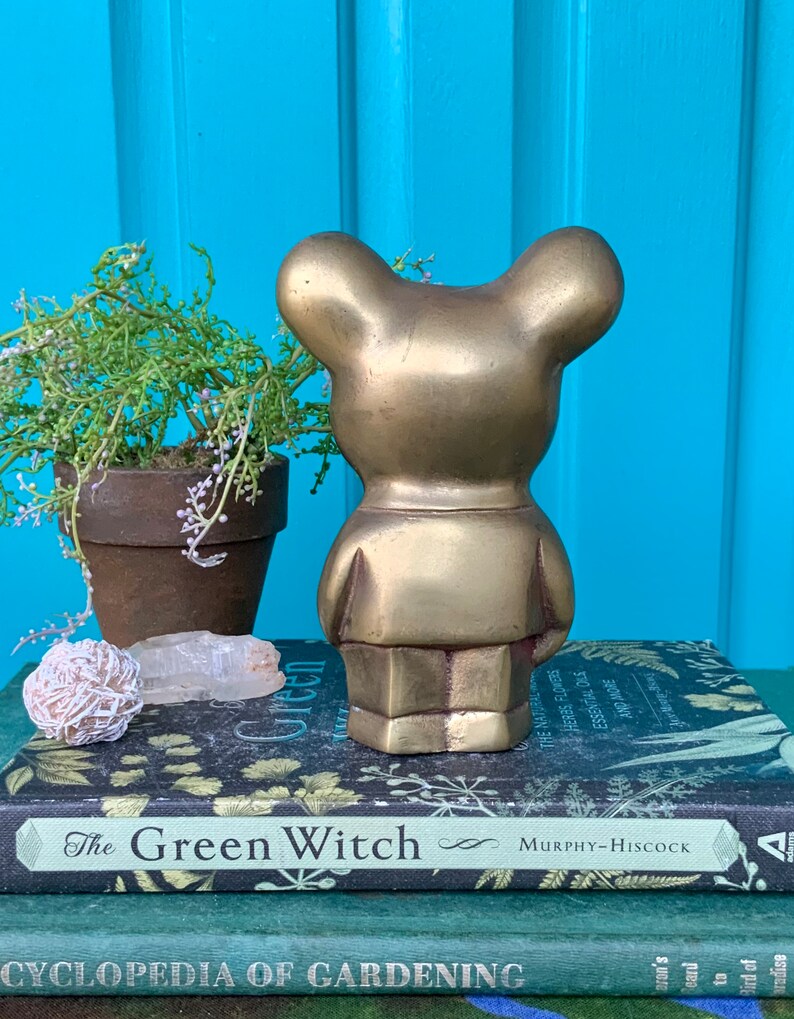 Op de afbeelding: Een gouden metalen berenbeeldje met een eenvoudig ontwerp. De beer staat op een stapel boeken met de titel "The Green Witch" zichtbaar.