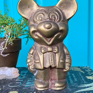 Op de afbeelding: Een gouden metalen beeldje van Mickey Mouse, gekleed in een pak en met een strikje. Het beeldje staat op een boek met een bloemenpatroon.
