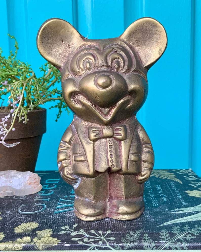 Op de afbeelding: Een gouden metalen beeldje van Mickey Mouse, gekleed in een pak en met een strikje. Het beeldje staat op een boek met een bloemenpatroon.