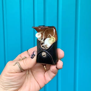 May include: A small, black leather case with a snap closure, holding vintage hearing aids. The case is open, revealing the hearing aids with white and yellow components. The background is a bright turquoise blue.