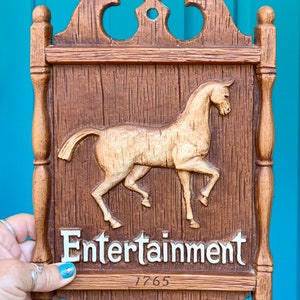 May include: A wooden sign with a carved horse and the word "Entertainment" in white letters. The sign has a brown woodgrain finish and a date of "1765" written below the word "Entertainment".