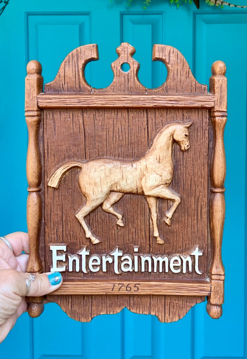 May include: A wooden sign with a carved horse and the word "Entertainment" in white letters. The sign has a brown woodgrain finish and a date of "1765" written below the word "Entertainment".