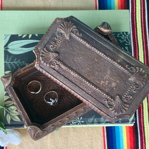 May include: A vintage wooden ring box with an ornate design. The box is open and has two silver rings inside.
